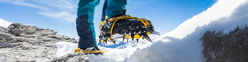 Person wearing Grivel crampons on a snowy and rocky mountain surface with a clear blue sky.