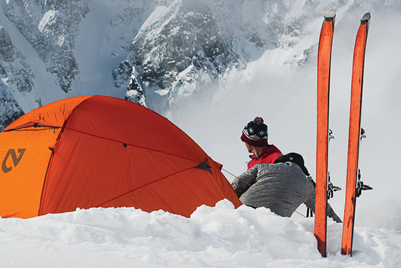 Person with skis and a Nemo alpine orange tent in a snowy mountain setting